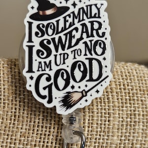 May include: A circular badge reel featuring a white acrylic topper with black text that reads I SOLEMNLY SWEAR I AM UP TO NO GOOD. The design includes a black witch hat, a small broomstick, and scattered stars. It has a clear plastic clip and metal hook.