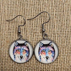 May include: A pair of silver-toned dangle earrings. Each earring features a round, white pendant with a watercolor-style wolf portrait in shades of blue, pink, and purple. The wolf has yellow eyes and a black nose.