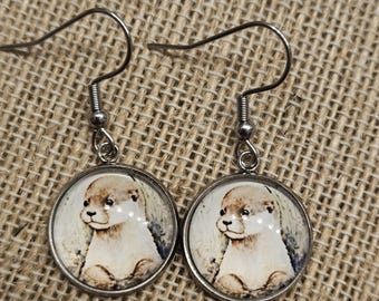 May include: A pair of dangle earrings featuring circular silver-toned metal settings. Each earring contains a round, glass-covered insert displaying a detailed, light-colored illustration of an otter with dark eyes and a small nose on a cream background.