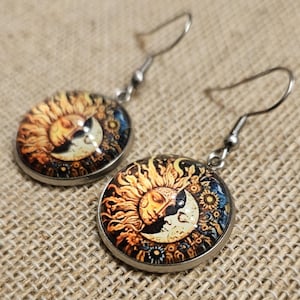 May include: A pair of silver-toned earrings featuring a celestial design. Each earring showcases a sun and moon motif with warm orange, yellow, and cream tones, set against a dark blue background. The earrings have a hook closure.