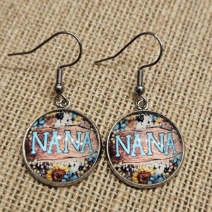 May include: A pair of round dangle earrings with the word "NANA" in light blue lettering. The earrings feature a rustic wood-like background with floral and butterfly accents, and silver-colored ear wires.