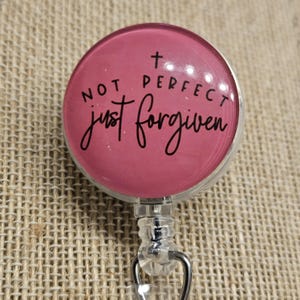 May include: A round, pink badge reel with the words "NOT PERFECT just forgiven" in black script. A small cross is above the word "perfect". The reel has a clear plastic body and a metal clip.