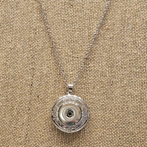May include: A silver-colored necklace with a round pendant. The pendant has a decorative floral design around the edge and a snap-in center. The necklace chain is also silver and hangs on a neutral-colored background.