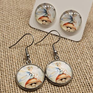May include: A set of fairy-themed earrings and matching stud earrings. The earrings feature a round, silver-toned setting with a glass dome. The image inside shows a fairy sitting on a mushroom. The fairy has blue wings and is wearing a blue dress.