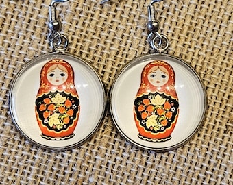 May include: Round silver earrings featuring a Russian nesting doll design. The dolls have a red headscarf, black dress, and orange floral details. The earrings hang from silver hooks.