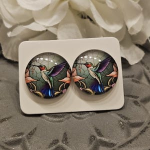 May include: A pair of round glass earrings with a stained glass design of a hummingbird in flight. The hummingbird is green, blue, and red with a long beak. The background is a green and brown pattern with pink flowers.
