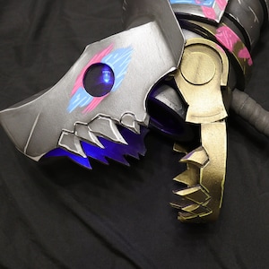 Könnte beinhalten: Nahaufnahme einer Requisitenwaffe mit mechanischem Design. Die Waffe hat einen silbernen Kopf mit blauen und rosa Akzenten, eine goldfarbene Klaue und einen schwarzen Griff. Das Design umfasst scharfe Kanten und ein blaues Leuchten.