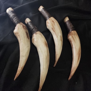 Puede incluir: Cuatro objetos curvos, blanquecinos, con puntas marrones y mangos negros envueltos, están dispuestos sobre una superficie negra. Los objetos tienen una apariencia texturizada, posiblemente parecida a dientes o garras. La estética general sugiere un tema primitivo o de fantasía.