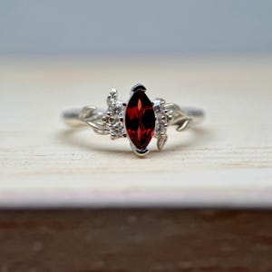 May include: A silver ring with a marquise-cut, deep red gemstone. The central stone is surrounded by smaller, clear stones and intricate, leaf-shaped embellishments. The ring is displayed on a light wooden surface.