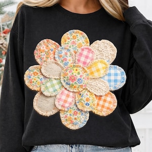 Könnte beinhalten: Schwarzes Sweatshirt mit einem großen, farbenfrohen Blumendesign. Die Blume besteht aus verschiedenen Stoffflicken mit floralen, Vichy- und Spitzenmustern. Die Flicken sind in den Farbtönen Rosa, Gelb, Blau und Orange gehalten.