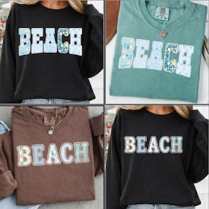 May include: Four long-sleeved sweatshirts in black, teal, brown, and black. Each shirt features the word "BEACH" in a white, blue, and floral design. The shirts are casual and suitable for beach or summer wear.