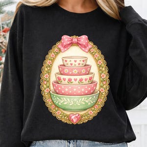 May include: Black sweatshirt featuring a decorative design of stacked bowls. The bowls are pink and green, adorned with floral and heart patterns. A pink bow and heart-shaped gem complete the design, set within a gold-colored oval frame.