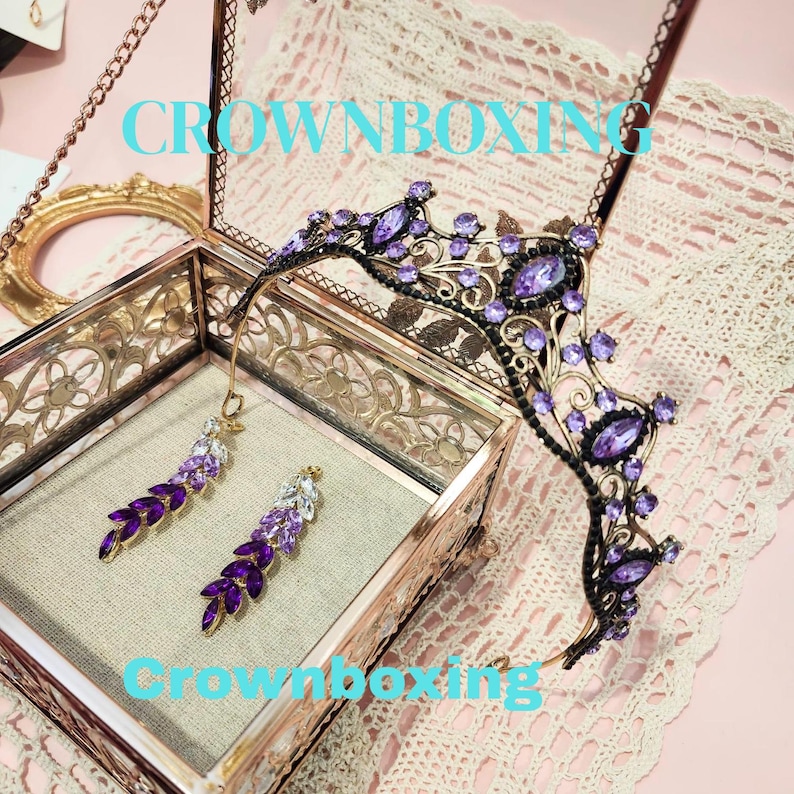 Puede incluir: Una corona decorativa y pendientes a juego con piedras preciosas moradas. La corona es negra con detalles ornamentados y los pendientes tienen un dise&ntilde;o de hojas colgantes. Las joyas se exhiben en una caja de cristal decorativa con el texto "Crownboxing".