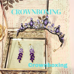 Puede incluir: Una tiara negra y morada y un juego de pendientes a juego exhibidos en una caja de cristal. La tiara presenta piedras preciosas moradas y detalles de hojas. Los pendientes son una cascada de cristales morados y transparentes. El texto "CROWNBOXING" es visible.