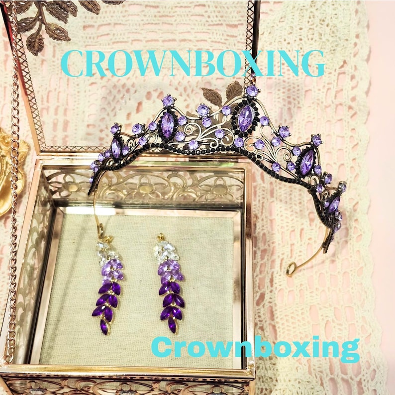 Puede incluir: Una tiara negra y morada y un juego de pendientes a juego exhibidos en una caja de cristal. La tiara presenta piedras preciosas moradas y detalles de hojas. Los pendientes son una cascada de cristales morados y transparentes. El texto "CROWNBOXING" es visible.