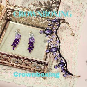 Puede incluir: Un conjunto de joyas decorativas con una tiara dorada y pendientes a juego. La tiara y los pendientes est&aacute;n adornados con cristales morados y transparentes. Los pendientes se exhiben dentro de una caja dorada decorativa. El texto "Crownboxing" es visible.