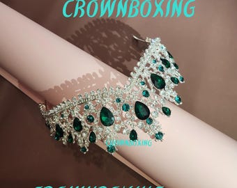 May include: A silver tiara with a cascading design of clear and emerald green rhinestones. The tiara is on a pink surface with the text "CROWNBOXING" on the surface.