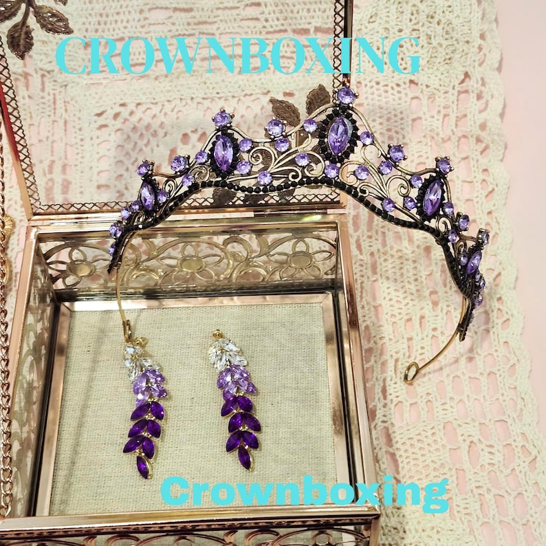 Puede incluir: Una corona ornamentada y pendientes a juego con piedras preciosas moradas y transparentes. La corona se exhibe sobre una caja de cristal decorativa. Los pendientes tienen un dise&ntilde;o de hojas. El texto "CROWNBOXING" es visible.