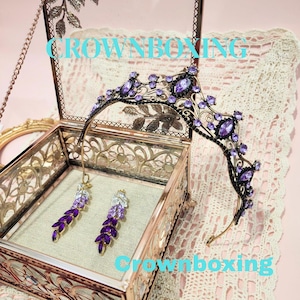 Puede incluir: Una tiara negra y morada con pendientes a juego, exhibida en una caja de cristal decorativa. La tiara y los pendientes est&aacute;n adornados con piedras preciosas moradas. El texto "CROWNBOXING" es visible en la imagen.