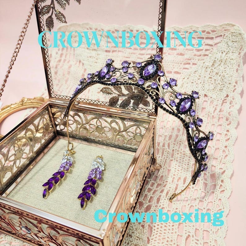 Puede incluir: Una tiara negra y morada con pendientes a juego, exhibida en una caja de cristal decorativa. La tiara y los pendientes est&aacute;n adornados con piedras preciosas moradas. El texto "CROWNBOXING" es visible en la imagen.