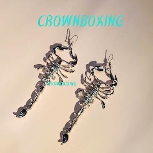 May include: A pair of silver scorpion earrings with dangling tails. The earrings are detailed with rhinestones and have a hook closure.