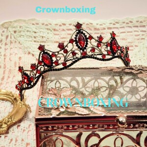 May include: A black and red jeweled crown with the text "Crownboxing" in light blue. The crown features red gemstones and an ornate design. The crown is displayed on a decorative box.