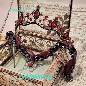 Op de afbeelding: Een zwarte en rode sierlijke kroon en ketting set in een decoratieve glazen doos. De kroon heeft rode edelstenen en sierlijk metaalwerk. De ketting heeft een ketting en rode stoffen accenten, met een hanger. De doos is goudkleurig.