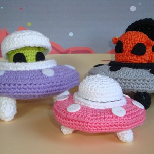 Puede incluir: Tres juguetes de OVNI de crochet. Uno es morado con un extraterrestre verde en su interior, otro es rosa con botones blancos y el último es gris con un extraterrestre naranja en su interior.