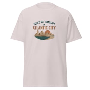 May include: A light pink short-sleeve t-shirt featuring a graphic print on the chest. The design displays a city skyline, a pier, and a sun, with the text "MEET ME TONIGHT IN ATLANTIC CITY" in dark teal and orange lettering above the illustration.