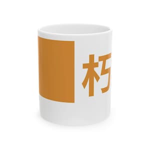Puede incluir: Taza de cerámica blanca con un gran cuadrado naranja y caracteres japoneses naranjas. La taza es cilíndrica y tiene un asa. El diseño es simple y moderno, adecuado para café, té u otras bebidas.
