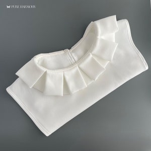 Pode incluir: Colete de tecido branco com uma borda franjada. O colete está dobrado ao meio e tem uma costura no centro.