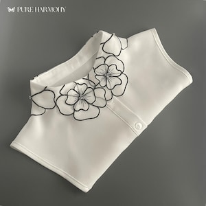 Puede incluir: Un cuello desmontable blanco con un diseño floral. El cuello presenta contornos negros de flores con cierre de botón. El texto "PURE HARMONY" es visible en la esquina superior izquierda.