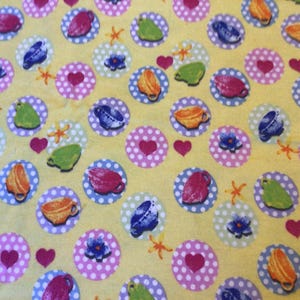 May include: Yellow fabric with a repeating pattern of colorful teacups, hearts, flowers, and starfish. The teacups are in shades of red, green, blue, and orange, set within white polka-dotted circles.