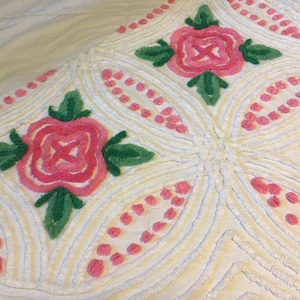 May include: Close-up of a cream-colored, tufted textile with a floral pattern. The design features pink roses with green leaves and pink dotted accents. The texture appears soft and plush, with a raised, looped pile.