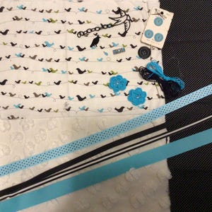 May include: A collection of craft supplies including fabric with a bird pattern, blue crochet flowers, ribbons, buttons, and thread. The fabric features small birds in various colors. Ribbons are black, white, and blue. Buttons are blue and black.