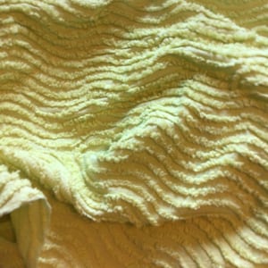 May include: Close-up of a soft, textured, light green fabric with a wavy, ridged pattern. The fabric appears plush and has a slightly uneven surface, creating shadows and highlights. The overall texture suggests a towel or blanket.