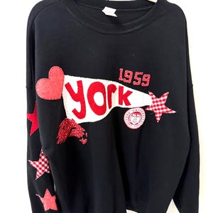 May include: Black sweatshirt with red and white appliqués. Features a red heart, stars, and the word "York" in red on a white banner. Includes the year "1959" and a circular emblem. The stars have gingham and glitter details.