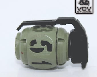 May include: A green and black plastic replica of a military grenade with a handle on top. The grenade has a face-like design with holes in the shape of eyes and a mouth.