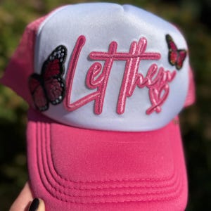 Op de afbeelding: Roze truckerpet met een wit voorpaneel. De pet heeft de tekst "Let them" in roze letters, samen met twee vlinderpatches. De pet heeft een roze klep en een mesh achterkant.