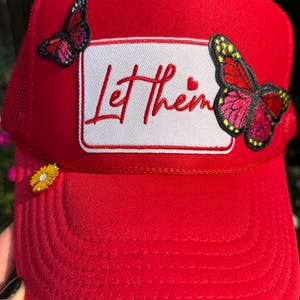 May include: A bright red trucker hat with a white rectangular patch featuring the embroidered phrase "Let Them" in red script. Two butterfly patches with red, pink, and yellow details adorn the hat, along with a small yellow flower.