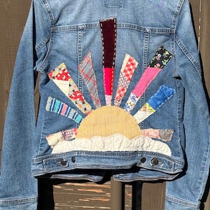 Puede incluir: Chaqueta vaquera azul claro con un diseño de sol de patchwork colorido en la espalda. El sol presenta un centro amarillo, nubes blancas y rayos en varias telas estampadas. La chaqueta tiene un frente abotonado clásico y bolsillos.