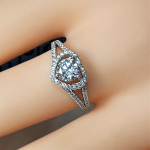 May include: A silver ring with a heart-shaped halo setting and a round, clear center stone. The ring is set with smaller clear stones around the center stone and the band.