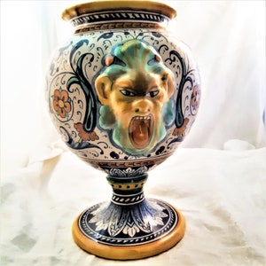 May include: A ceramic vase with a yellow, blue, and green floral design. The vase has a round body and a tall, slender base. A grotesque face with a wide open mouth is painted on the front of the vase.