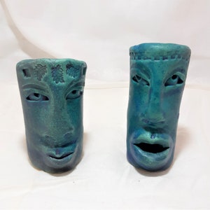 May include: Two teal blue ceramic face planters with carved details. One planter has a leaf design on the forehead and the other has a geometric design.