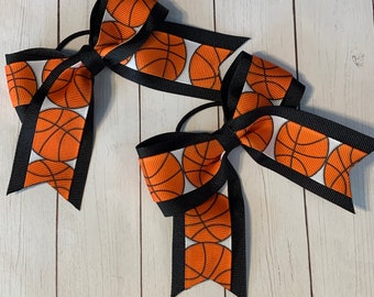 May include: Two black and white hair bows with orange basketball designs. The bows are tied together with a black elastic band.