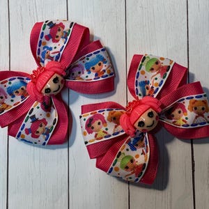 May include: Three pink hair bows with a cartoon doll design. The bows are made of grosgrain ribbon with a repeating pattern of dolls in various colors. Each bow has a pink doll face in the center.