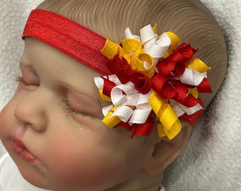 May include: A red elastic headband adorned with a multi-colored bow. The bow features red, white, and yellow ribbons arranged in a spiral design. The headband is worn on a doll.