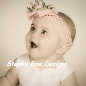 May include: A baby wearing a pink lace headband with a large bow adorned with flowers and ribbons. The baby is looking up with an open mouth, and the text "Stephs Bow Designs" is visible.