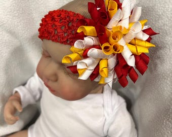 May include: A red, white, and yellow bow headband for a baby. The headband is made of crocheted elastic and features a large, fluffy bow.