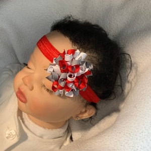 Könnte beinhalten: Ein rotes Stirnband mit einer grauen, weißen und roten Schleife. Die Schleife ist aus Band und zu einem Knoten gebunden. Das Stirnband wird von einem Baby getragen.